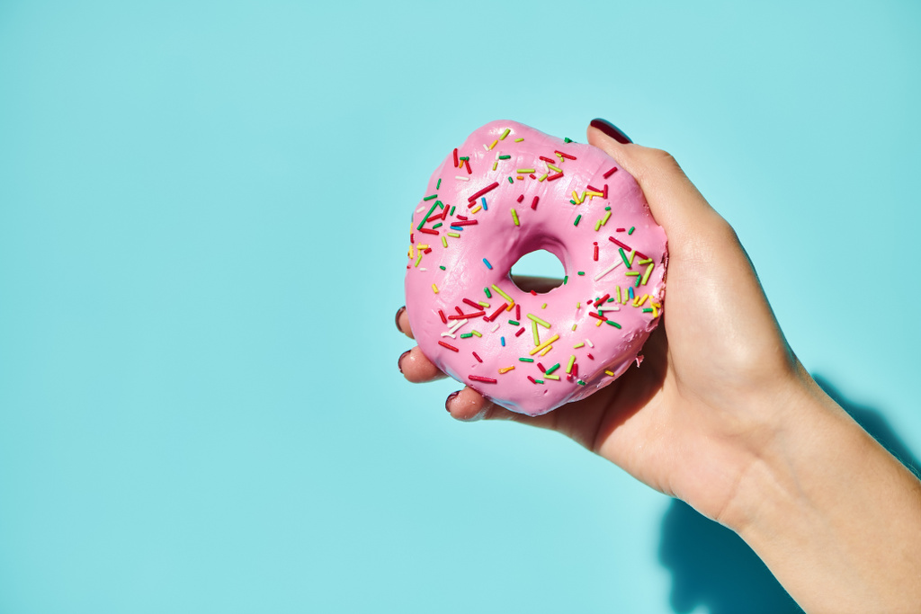 dulce y delicioso donut con glaseado rosa en la mano de una mujer desconocida sosteniéndolo sobre un fondo rosa - Foto, Imagen