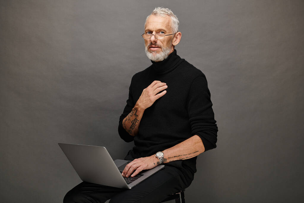 attraente uomo maturo in trendy dolcevita nero seduto sulla sedia con computer portatile e guardando la fotocamera - Foto, immagini