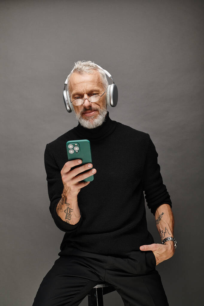 jóképű érett férfi modell szakállal és fejhallgatóval fekete garbóban nézi a telefont - Fotó, kép