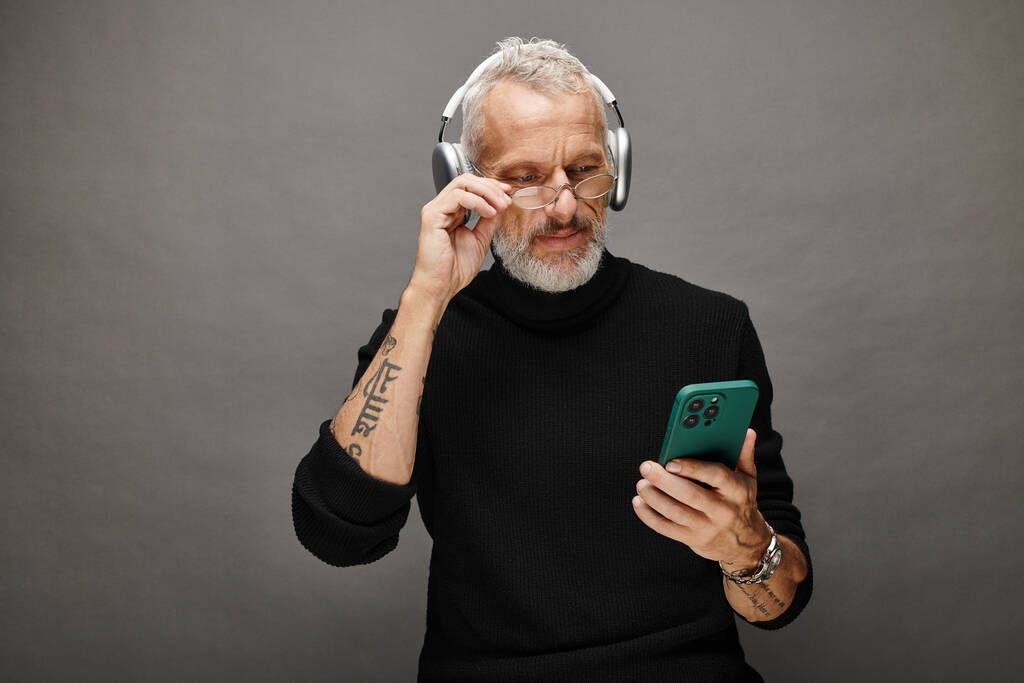 beau modèle masculin mature avec barbe et écouteurs en col roulé noir regardant le téléphone - Photo, image