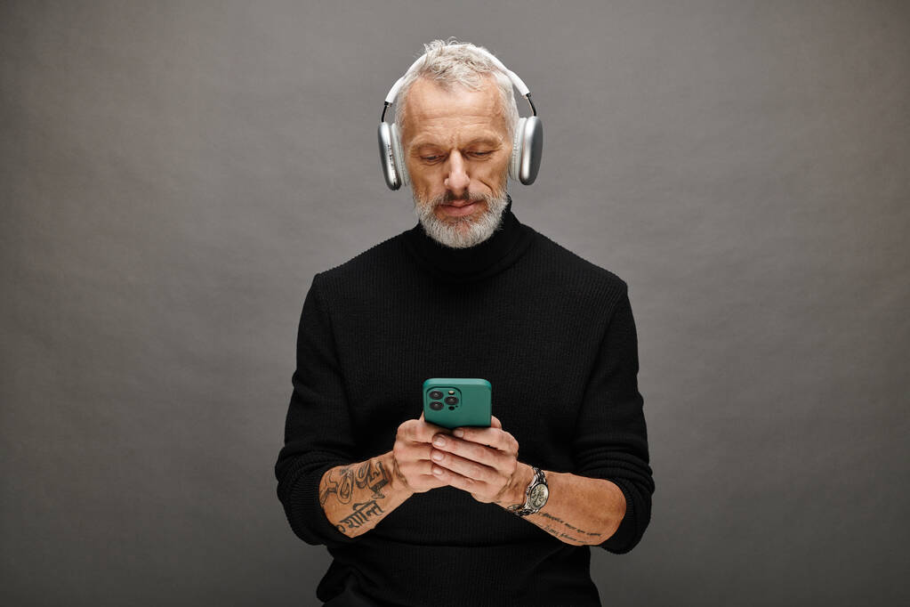 modelo masculino maduro de buen aspecto con barba y auriculares en cuello alto negro mirando el teléfono - Foto, Imagen