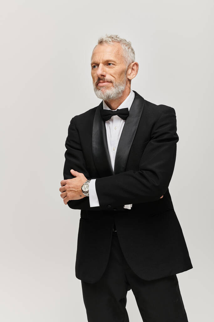 hombre maduro barbudo de buen aspecto en elegante esmoquin negro con pajarita posando y mirando hacia otro lado - Foto, Imagen