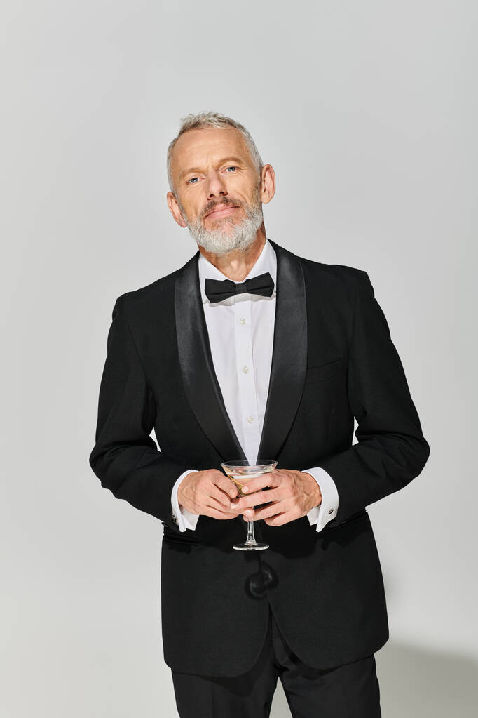 jóképű szakállas érett férfi modell elegáns szmokingban, martini üveget tartva és kamerába nézve - Fotó, kép