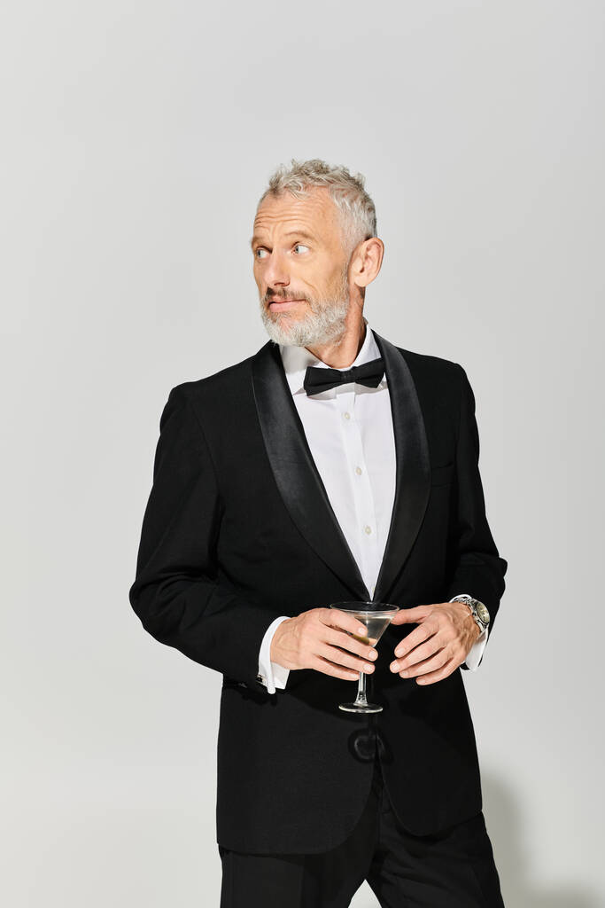 dobře vypadající dospělý muž v elegantním smokingu s plnovousem drží martini s olivami a dívá se jinam - Fotografie, Obrázek