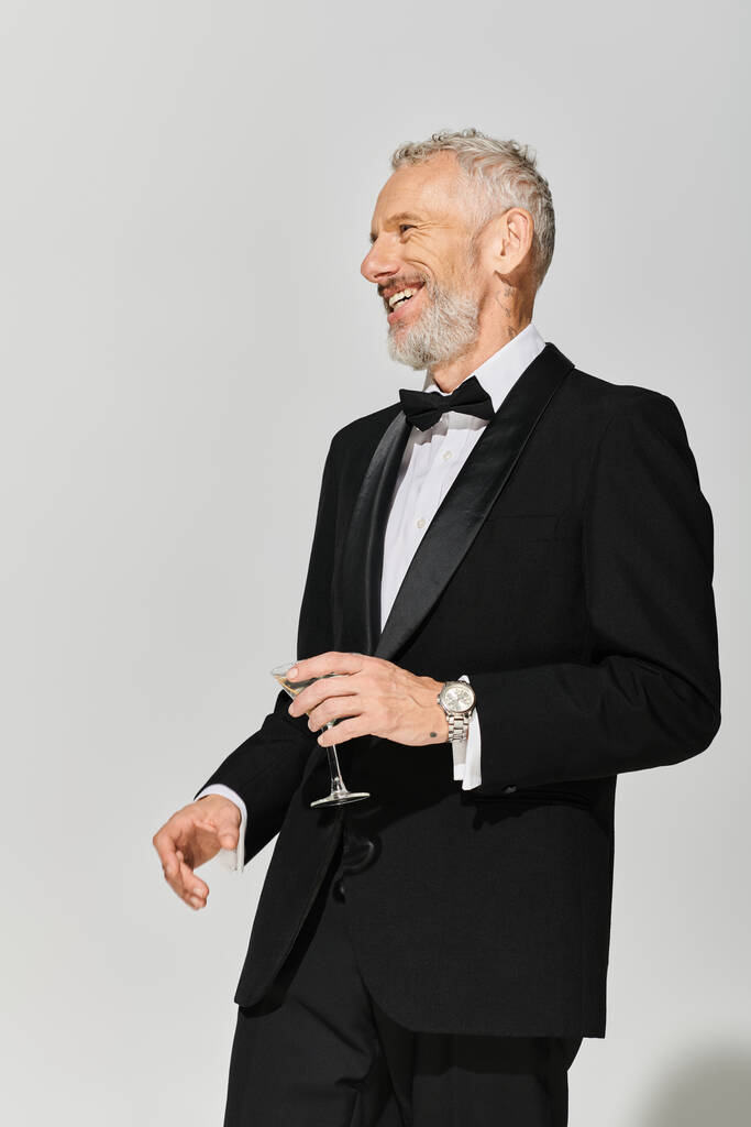 boldog, elegáns, érett férfi szakállal, debonair szmokingban, martinit tartva és vidáman mosolyogva. - Fotó, kép