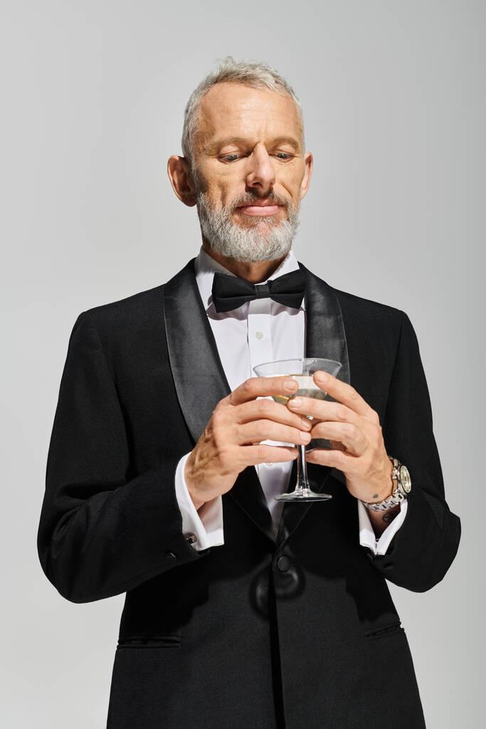 attraktiver bärtiger älterer Mann im eleganten Smoking mit Fliege, der ein Glas Martini mit Oliven hält - Foto, Bild