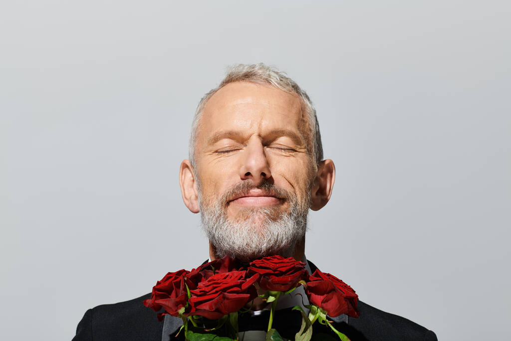 alegre hombre maduro de buen aspecto en esmoquin elegante con ramo de rosas rojas y sonriendo con los ojos cerrados - Foto, Imagen