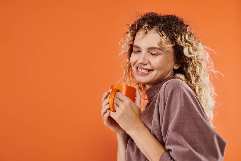elégedett nő göndör haj és csésze reggeli kávé mosolygó csukott szemmel narancs - Fotó, kép