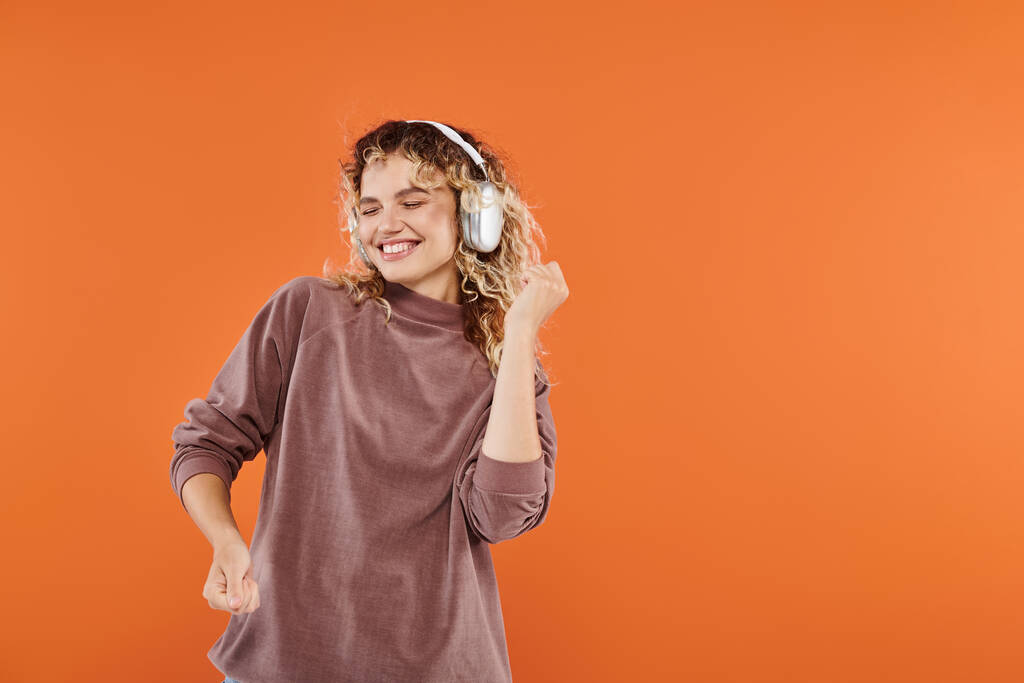 podekscytowana kobieta z falowane włosy taniec w słuchawkach bezprzewodowych i mocha kolor golf na pomarańczowy - Zdjęcie, obraz