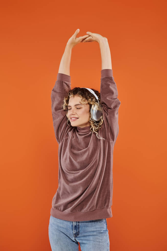 glad curly woman with closed eyes and raised hands listening music in wireless headphones on orange - Photo, Image
