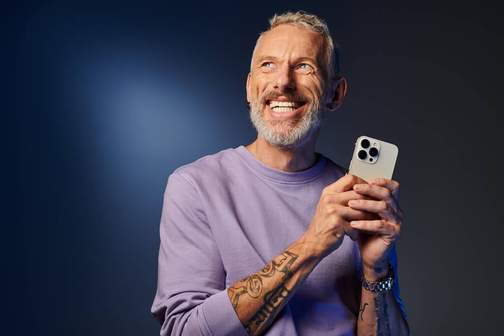 good looking joyous mature man with beard in fashionable sweatshirt holding phone and looking away - Photo, Image