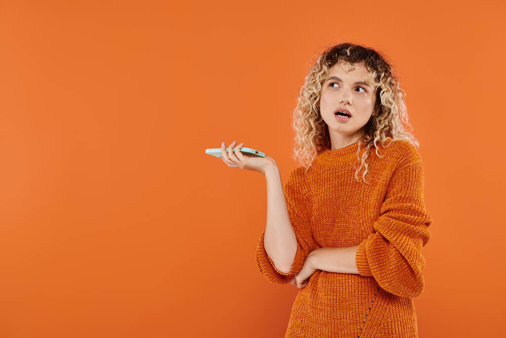 mujer rizada reflexiva en suéter brillante sosteniendo en el teléfono móvil y mirando hacia otro lado en el fondo naranja - Foto, Imagen