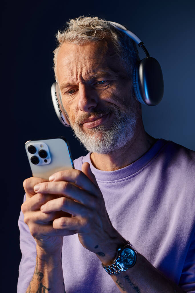 good looking bearded mature man in purple sweatshirt with headphones looking at his smartphone - Photo, Image