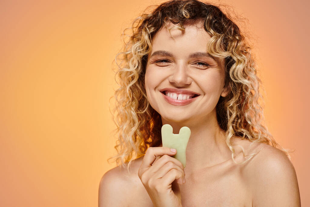 cheerful curly woman with perfect skin holding gua sha looking at camera on pastel backdrop - Photo, Image