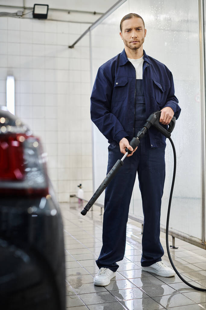 bonito profissional serviceman em azul uniforme segurando mangueira antes de usá-lo para lavar carro preto - Foto, Imagem