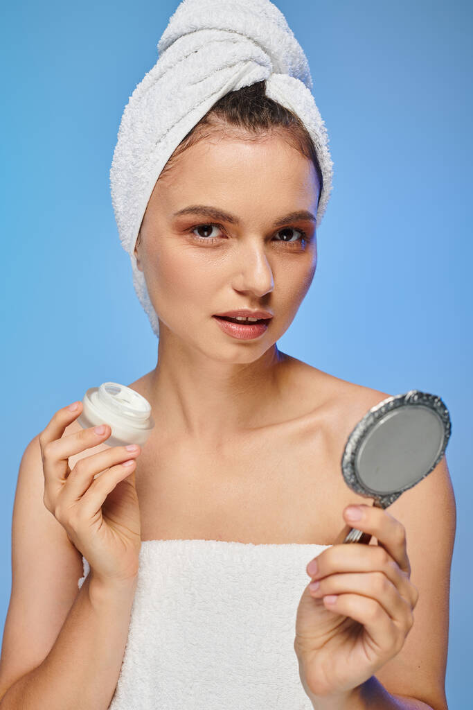 attraktive Frau mit Gesichtscreme und Handtuch auf dem Kopf, die vor blauem Hintergrund in die Kamera schaut, Selbsthilfe - Foto, Bild