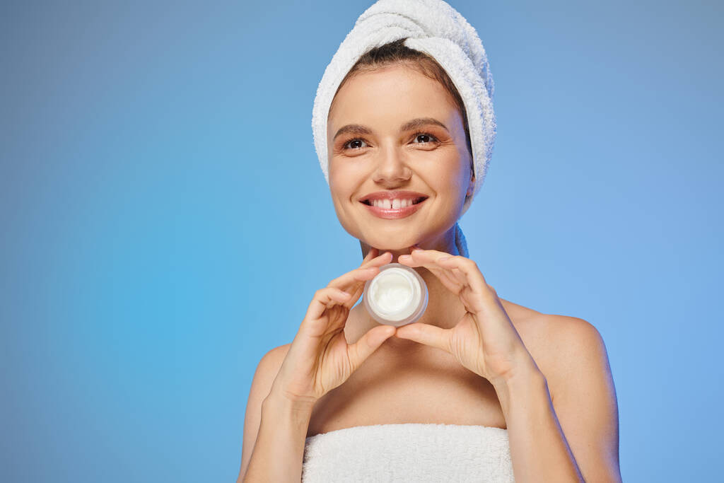 smiling model with towel on head holding jar of face cream on blue backdrop, wellness and beauty - Photo, Image