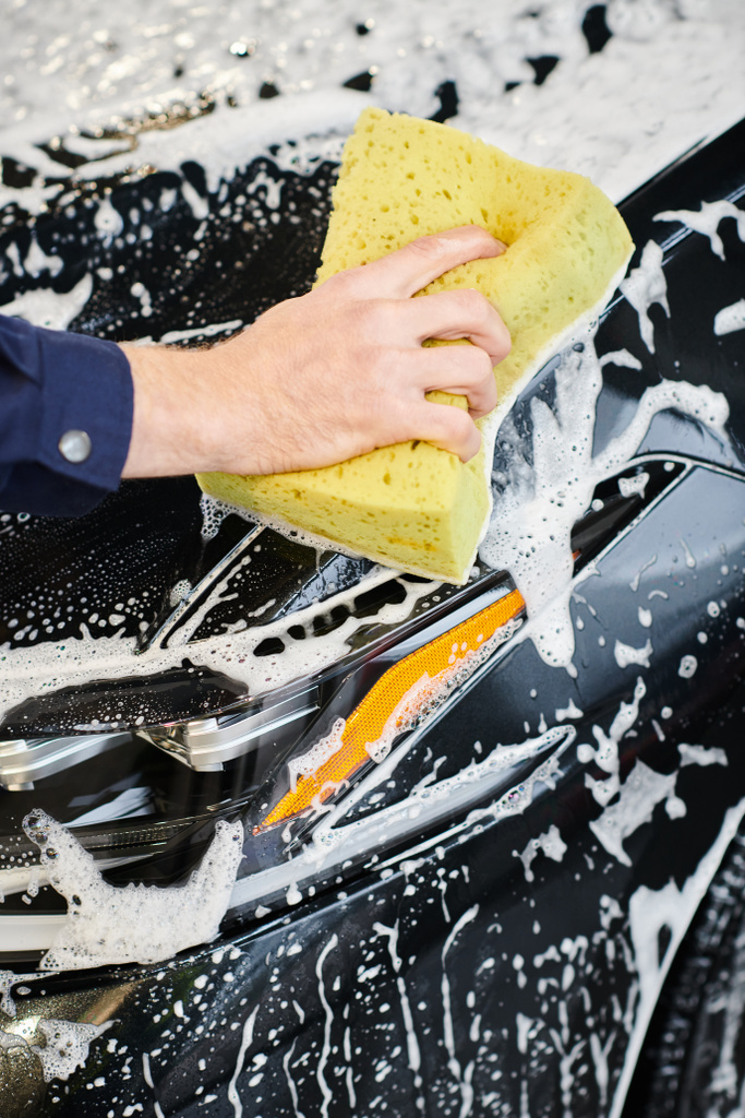 Siyah modern arabayı yıkamak için sabunlu sünger kullanan mavi üniformalı bir uzman. - Fotoğraf, Görsel