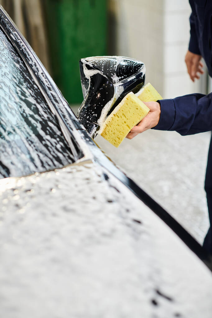 vue recadrée d'un spécialiste dévoué travaillant dur dans une voiture de lavage uniforme confortable avec une éponge savonneuse - Photo, image