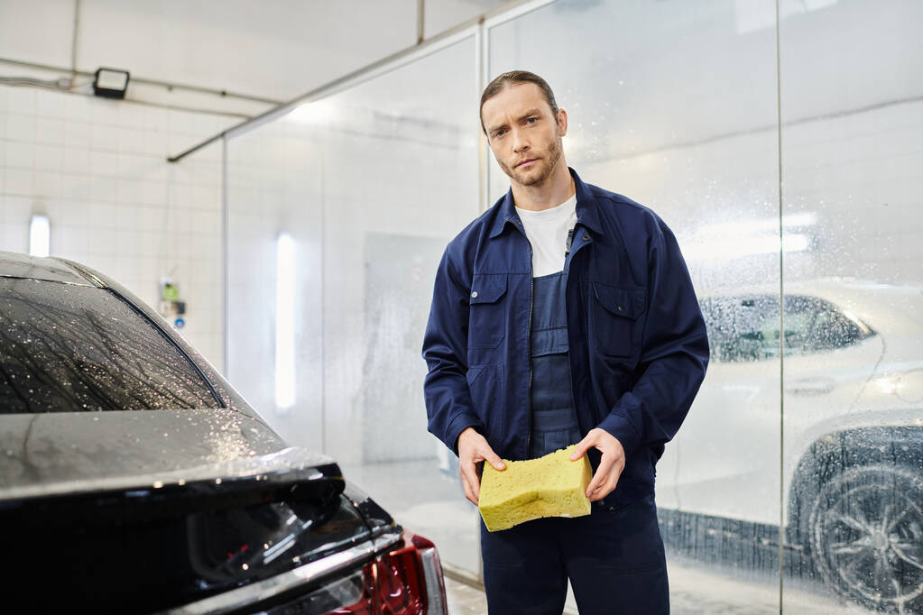 красивый профессиональный работник в синей форме держит мыльную губку и смотрит в камеру в гараже - Фото, изображение