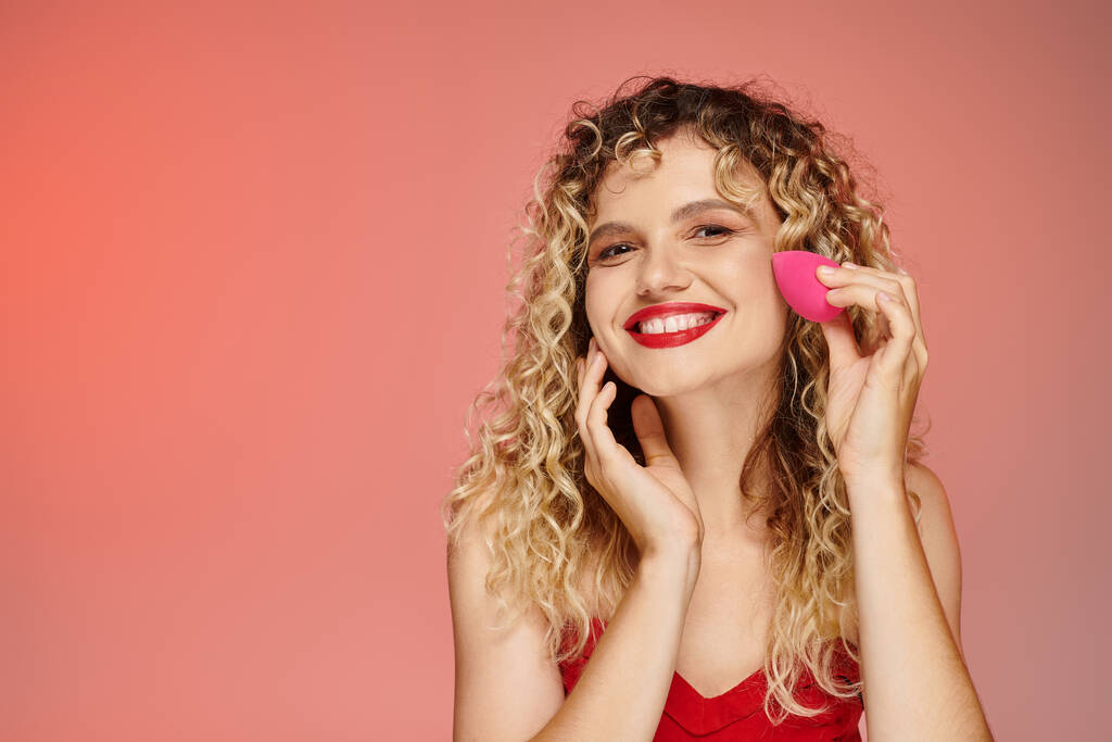 curly joyful model with red lips using face sponge on pink and yellow backdrop, elegant beauty - Photo, Image