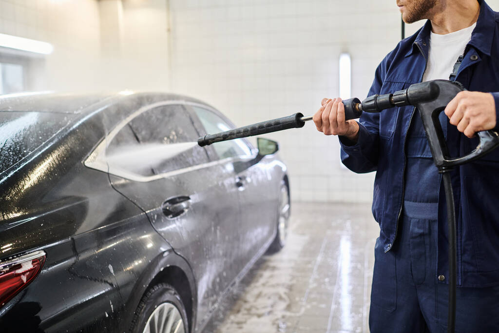 przycięty widok oddanego profesjonalnego pracownika w niebieskim mundurze za pomocą węża do mycia czarny nowoczesny samochód - Zdjęcie, obraz