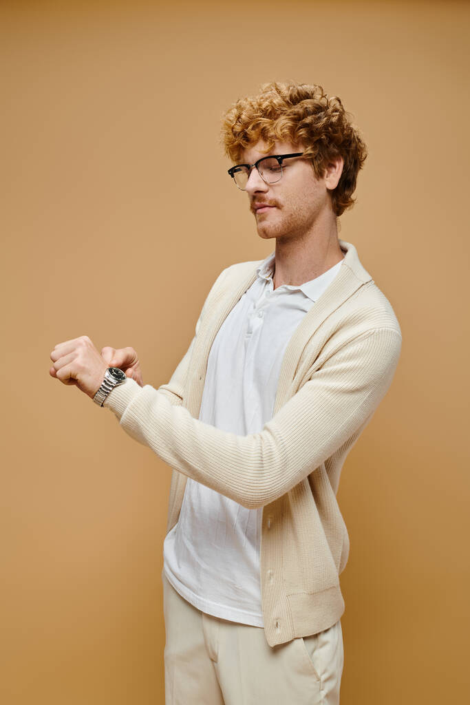 módní zrzka muž v brýlích při pohledu na hodinky na béžové pozadí, nadčasové staré peníze móda - Fotografie, Obrázek