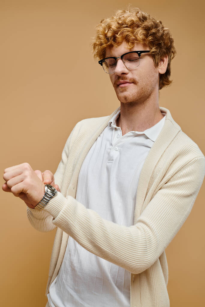 όμορφος νεαρός άνδρας με γυαλιά που κοιτάζει το ρολόι χειρός σε μπεζ, αριστοκρατικό παλιό στιλ χρήματος - Φωτογραφία, εικόνα
