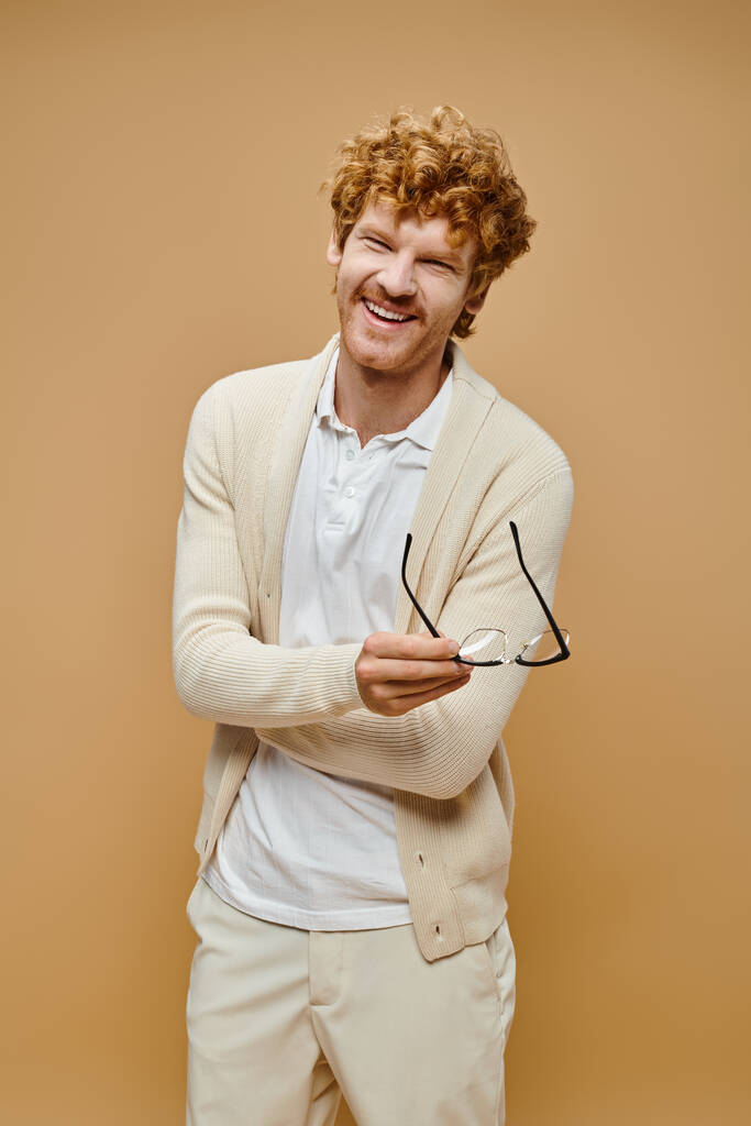 hombre pelirrojo excitado en ropa de color claro sosteniendo anteojos y riendo sobre fondo beige - Foto, imagen