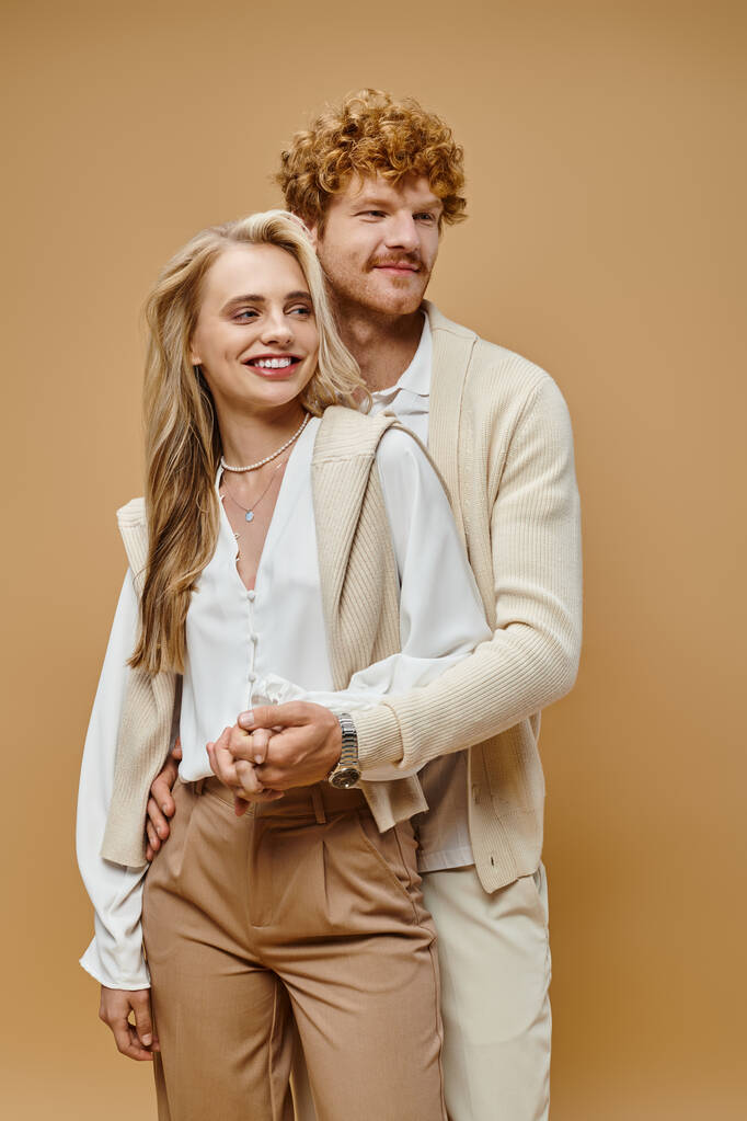 positive blonde Frau und rothaarige Mann in heller Kleidung Händchen haltend und wegschauend auf beige - Foto, Bild