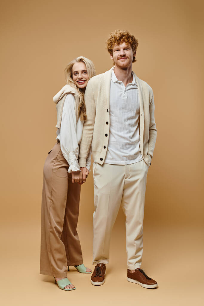 Moda kıyafetleri içinde, el ele tutuşmuş bej renkli kameraya bakan mutlu genç bir çift. - Fotoğraf, Görsel