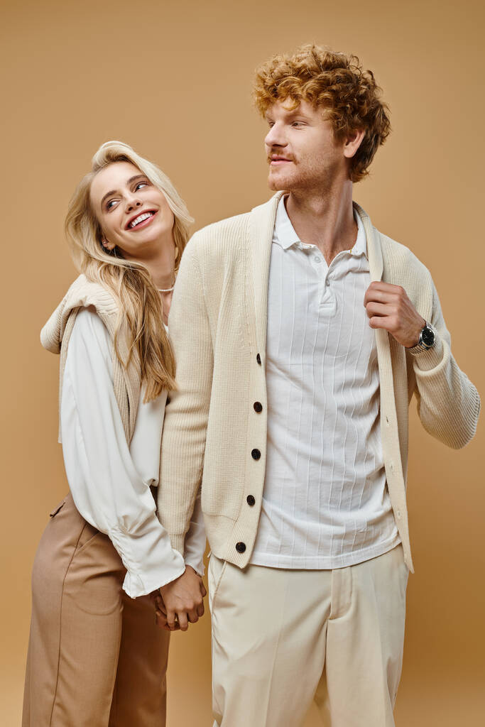 old money style, young cheerful couple in trendy outfit holding hands and looking away on beige - Photo, Image
