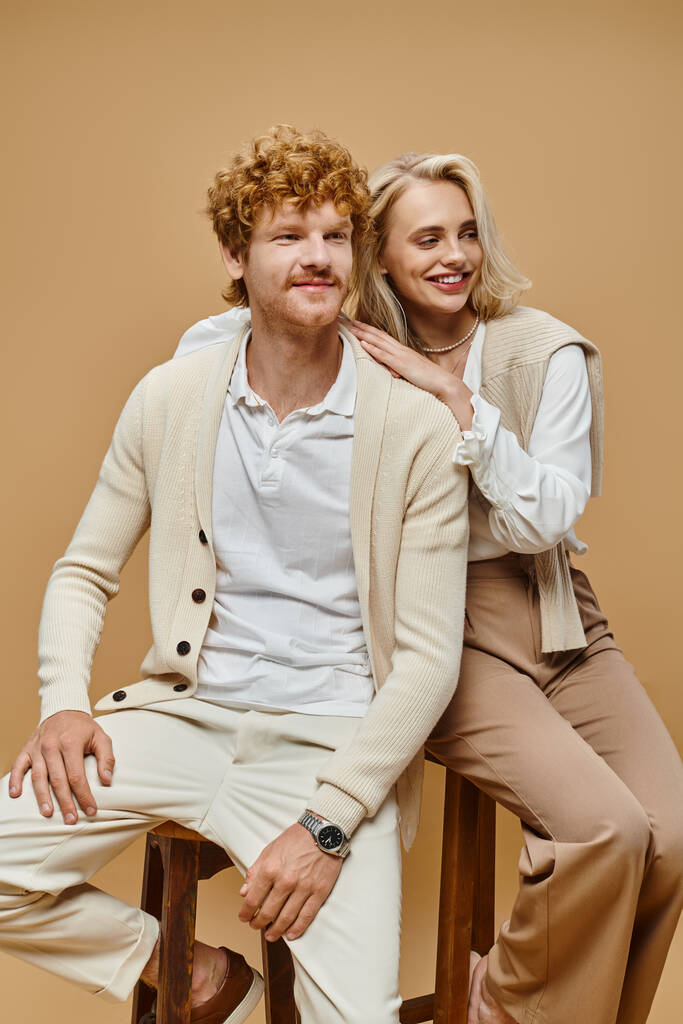 веселая молодая пара в модной одежде сидит на стульях и смотрит в сторону на бежевом фоне - Фото, изображение
