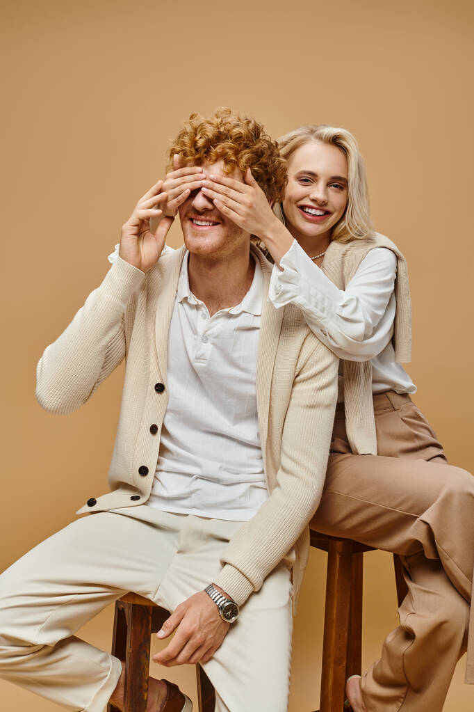 excité femme blonde assis sur la chaise et couvrant les yeux de rousse homme à la mode sur fond beige - Photo, image