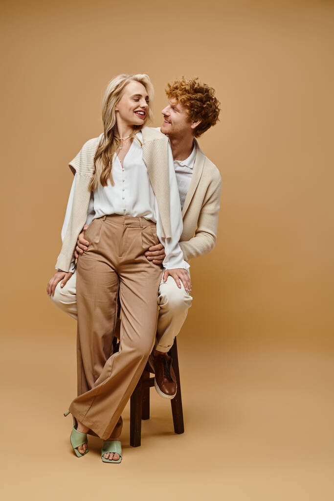 jolly redhead man sitting on chair near fashionable blonde woman on beige, timeless classic fashion - Photo, Image