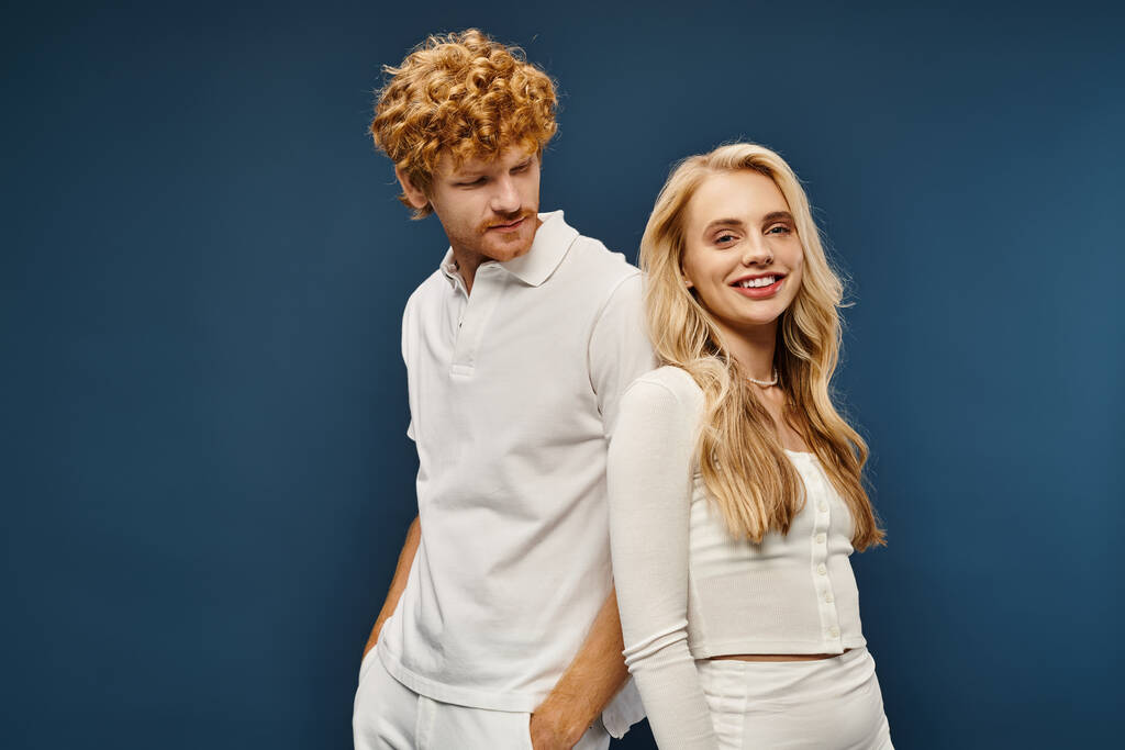 cheerful blonde woman in white trendy clothes standing back to back with redhead man on blue - Photo, Image