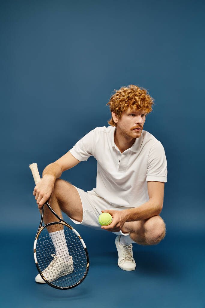 homme élégant en tenue blanche avec raquette de tennis et ballon assis sur des hanches regardant loin sur bleu - Photo, image