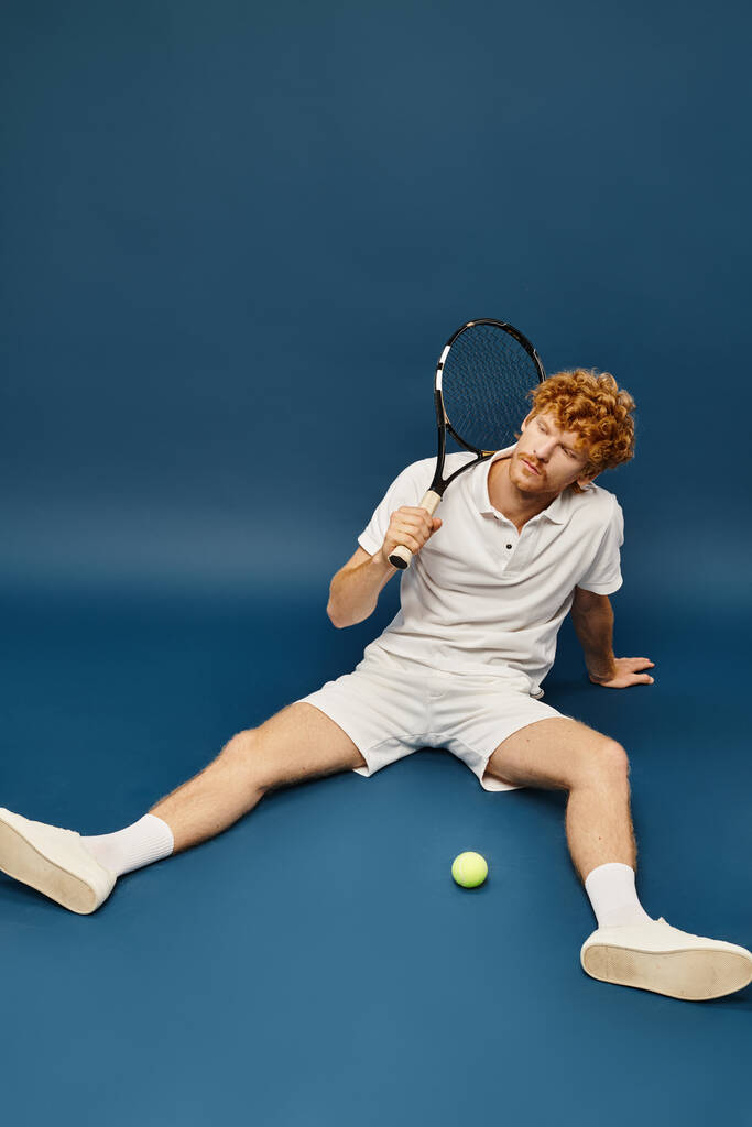 hombre pelirrojo deportivo en ropa blanca de moda con raqueta sentado cerca de la pelota de tenis sobre fondo azul - Foto, imagen