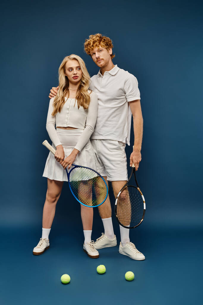 volledige lengte van trendy paar in wit tennis outfit poseren met tennisrackets in de buurt van ballen op blauw - Foto, afbeelding