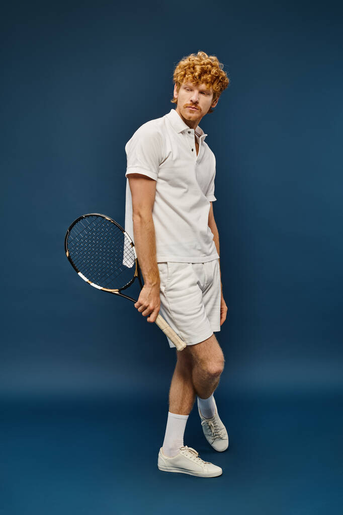 полная длина модного рыжеволосого человека в белом наряде с теннисной ракеткой, стоящей на синем фоне - Фото, изображение