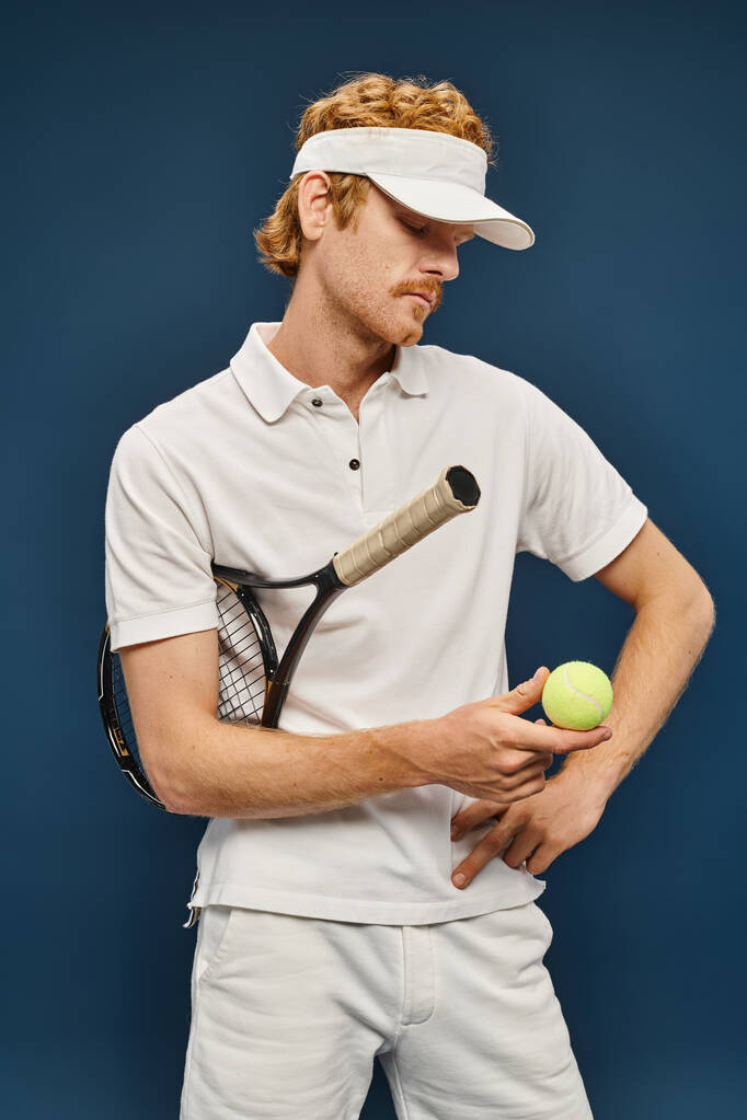 młody rudzielec w białych ubraniach i osłona przeciwsłoneczna trzymając rakietę i patrząc na piłkę tenisową na niebiesko - Zdjęcie, obraz