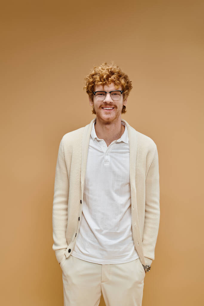 gelukkig roodharige man in bril en lichte kleding met handen in zakken kijkend naar camera op beige - Foto, afbeelding