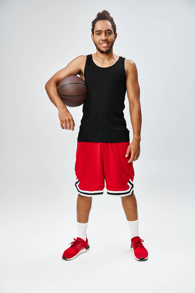pohledný veselý africký Američan ve sportovním oblečení drží basketbal a dívá se na kameru - Fotografie, Obrázek