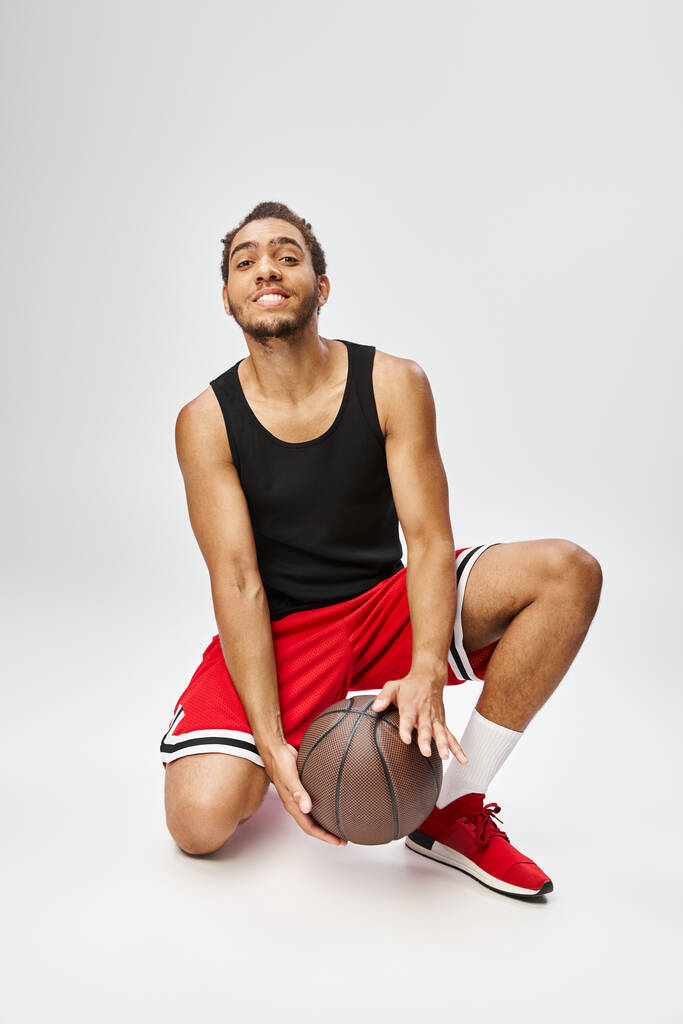 alegre afroamericano hombre posando con su baloncesto y sonriendo a la cámara sobre fondo gris - Foto, imagen