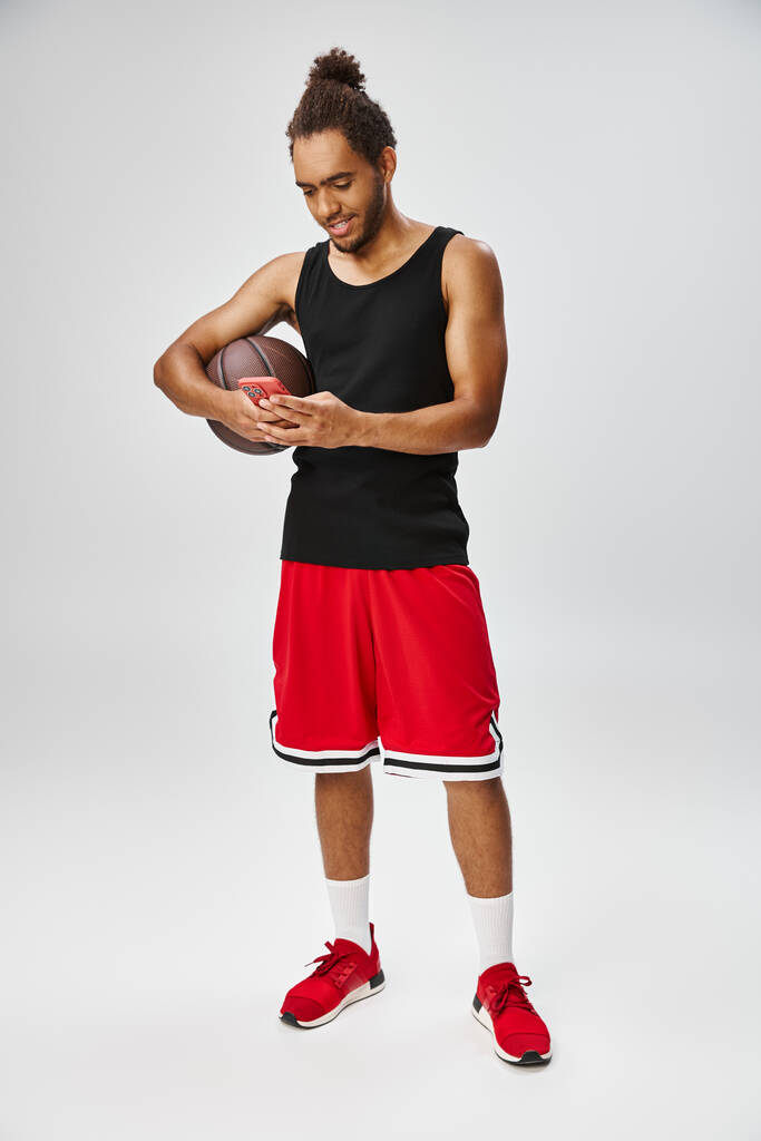vidám vonzó afro-amerikai sportoló gazdaság kosárlabda és nézi a telefon, online fogadás - Fotó, kép