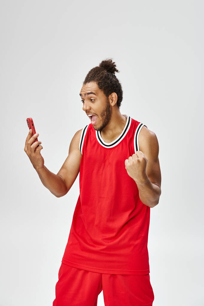 felice uomo afroamericano scioccato in abbigliamento sportivo tifo e guardando smartphone, scommesse online - Foto, immagini