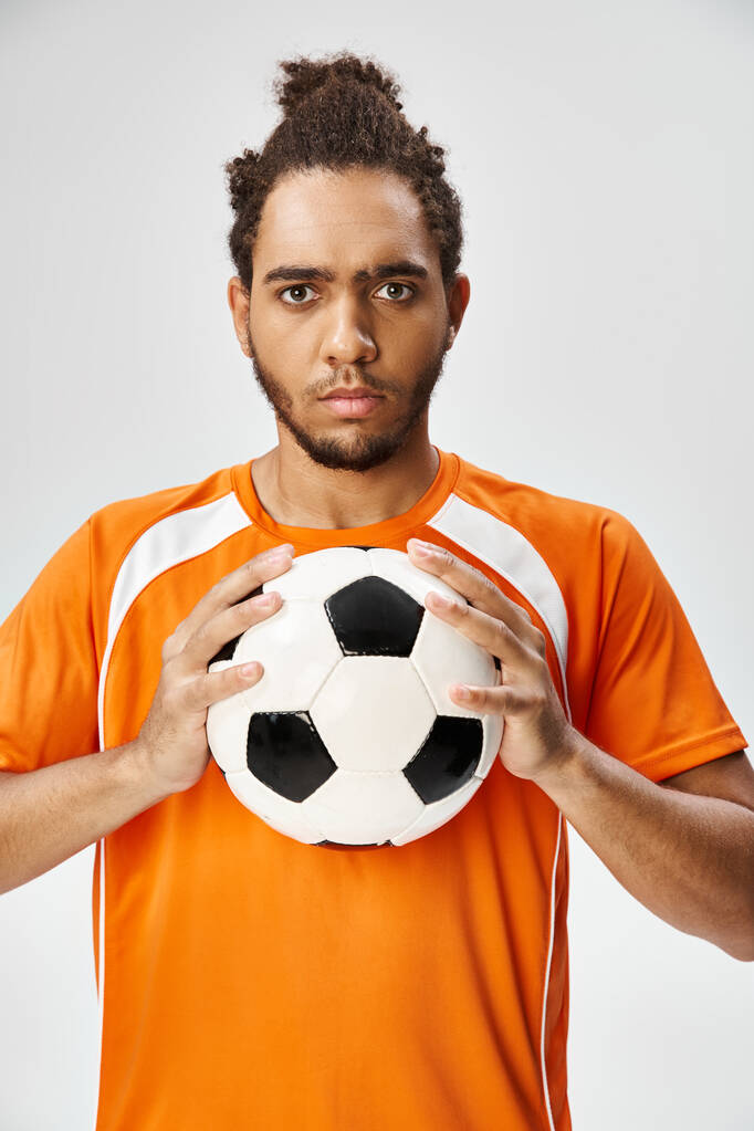 aantrekkelijke sportieve Afrikaanse Amerikaanse man poseren met voetbal en kijken naar de camera op grijze achtergrond - Foto, afbeelding