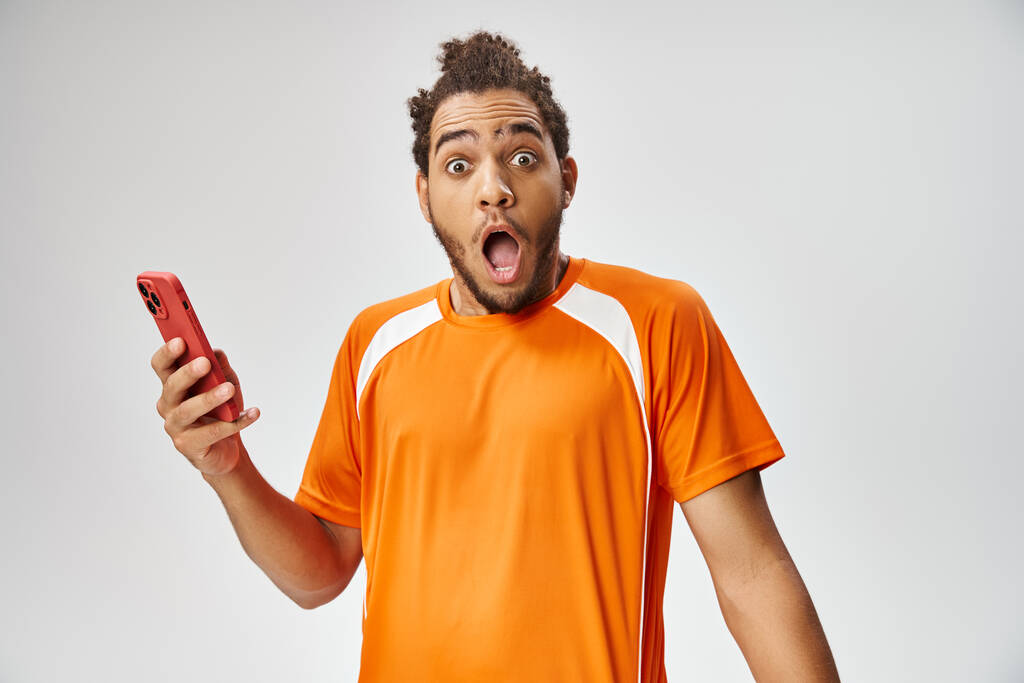 sorprendido hombre afroamericano en ropa deportiva celebración de teléfono y mirando a la cámara, apuestas en línea - Foto, imagen
