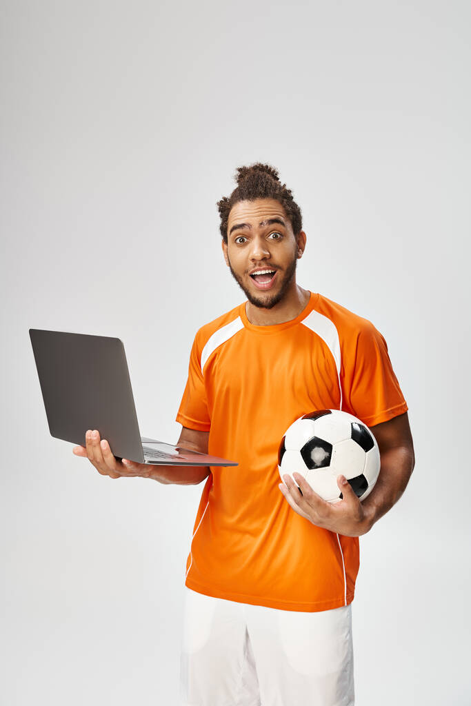 allegro uomo afroamericano in abbigliamento sportivo in possesso di calcio e laptop e guardando la fotocamera - Foto, immagini