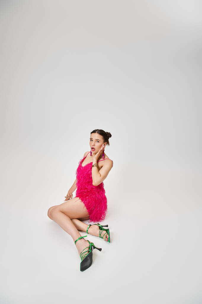 Playful girl in trendy pink dress and green shoes touching her cheek and sitting on grey background - Photo, Image
